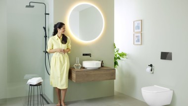 En kvinde i gul kjole står foran et mintgrønt badeværelse med badeværelsesmøbler og -porcelæn fra Geberit og sorte håndvaskarmaturer